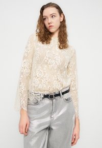 Top à manches longues en dentelle blanc cassé avec motif floral, associé à un jean argenté brillant taille haute et une ceinture noire à boucle ronde.