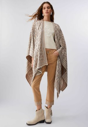 Donna che indossa uno scialle beige con motivo, maglione crema con applicazione floreale, pantaloni color tan con risvolto e stivaletti crema su sfondo bianco.