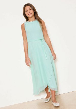 Mädchen steht in einem ärmellosen hellblauen Kleid mit Perlenverzierung am Ausschnitt und silbernen Ballettschuhen vor einer schlichten weißen Wand.