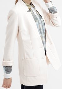 Light pink blazer with two front pockets, structured shape, and rolled sleeves, layered over a patterned shirt with blue and gray tones.