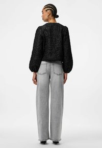 Svart teksturert bluse med volumsfylte ermer, kombinert med høytlivede, lys grå vide jeans. Bakfra, med to baklommer.