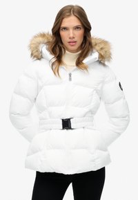 Chaqueta de plumas blanca con cintura ceñida, capucha con borde de piel, cierre de cremallera y textura acolchada. Presenta bolsillos laterales y detalle del logo en la manga.