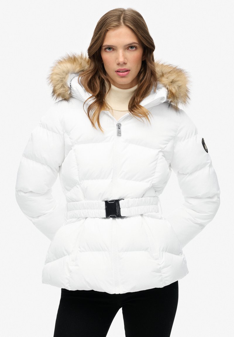 Chaqueta de plumas blanca con cintura ceñida, capucha con borde de piel, cierre de cremallera y textura acolchada. Presenta bolsillos laterales y detalle del logo en la manga.