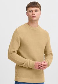 Beige gerippter Pullover mit rundem Halsausschnitt und langen Ärmeln, mit strukturierten Streifen und eng anliegenden Bündchen, kombiniert mit blauen Jeans.