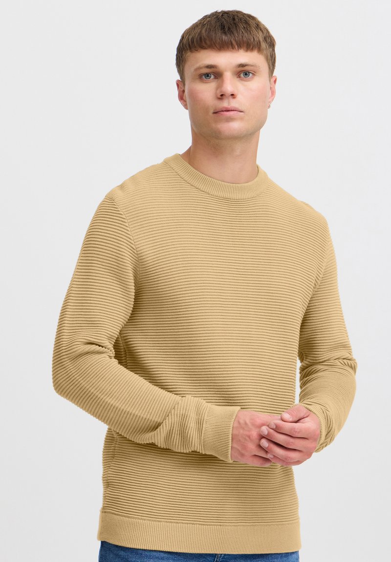 Beige gerippter Pullover mit rundem Halsausschnitt und langen Ärmeln, mit strukturierten Streifen und eng anliegenden Bündchen, kombiniert mit blauen Jeans.