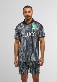 Mann in schwarzem und grauem gemustertem Sporttrikot und Shorts mit Logos, steht vor einem schlichten hellen Hintergrund.