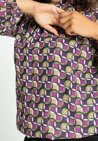 Personne portant un haut à manches longues avec un motif géométrique en violet, vert, beige et noir, mains levées près du visage.
