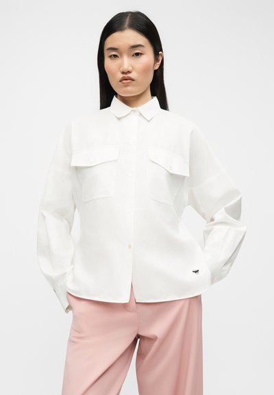 Γυναίκα με μακριά σκούρα μαλλιά, που φοράει λευκό πουκάμισο με κουμπιά και τσέπες στο στήθος και ροζ παντελόνι, στέκεται με τα χέρια στις τσέπες.