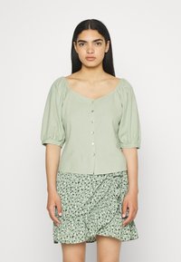 Blusa verde claro com mangas bufantes e decote quadrado, com fecho de botão, usada com uma saia rodada verde com padrão.