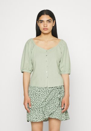Blusa verde claro com mangas bufantes e decote quadrado, com fecho de botão, usada com uma saia rodada verde com padrão.