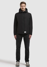 Sort zip-up jakke med hætte, der har en glat tekstur og justerbare snore, parret med mørke jeans og sneakers.