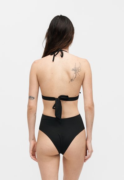 Γυναίκα με μακριά σκουρόχρωμα μαλλιά, που φοράει μαύρο μαγιό τύπου halter, εμφανίζεται από πίσω, με τατουάζ στην άνω πλάτη και στο αριστερό μπράτσο.