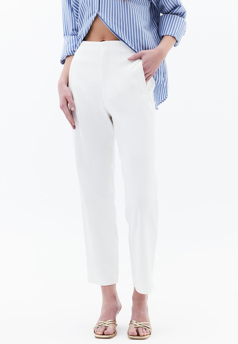Pantalons blancs ajustés avec une taille haute et des poches latérales. Tissu lisse et longueur raccourcie. Associés à une chemise rayée bleue.
