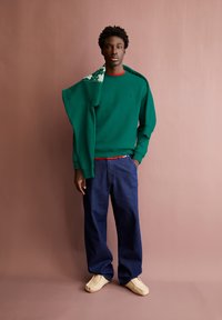 Felpa verde con scollatura rotonda, indossata sopra una maglietta rossa. Jeans blu larghi e scarpe beige senza lacci. Design semplice e casual.