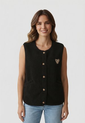 Donna che indossa un gilet in pile nero senza maniche con bottoni e toppa a forma di cuore con stampa leopardata, jeans azzurri, sorridente su sfondo neutro.