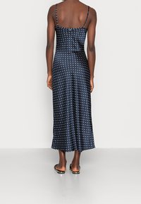 Femme de dos portant une robe en satin bleu marine à pois blancs et des sandales plates noires, sur un fond uni.