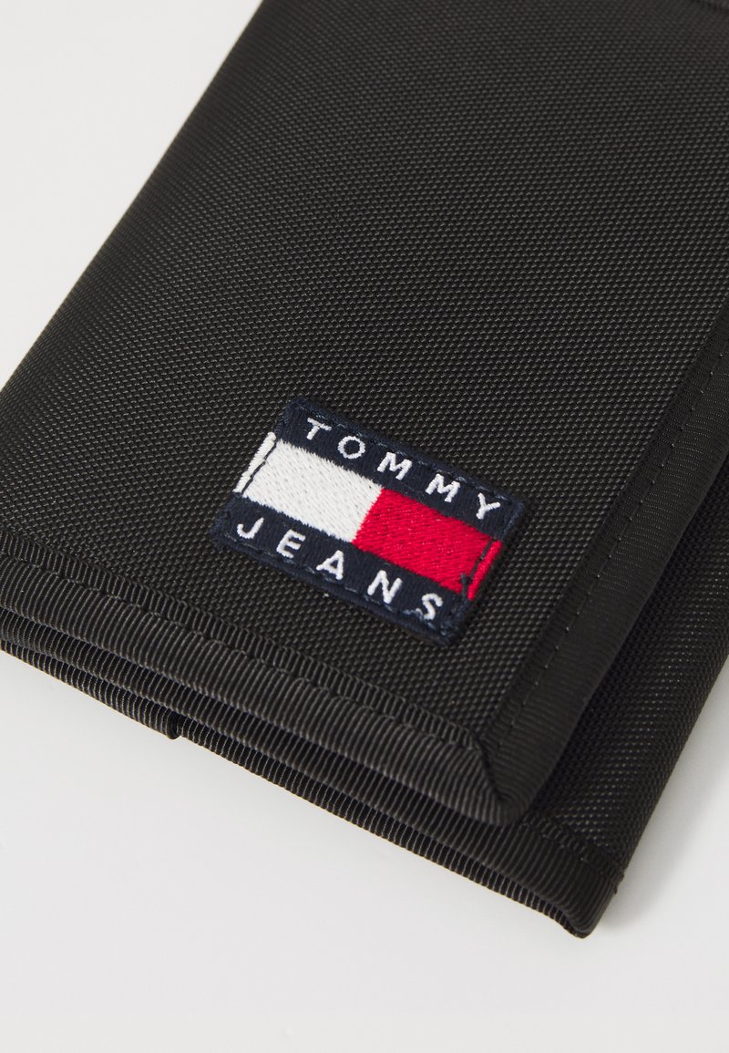 Cartera de tela negra con un logo bordado rectangular en azul marino, rojo y blanco que dice "TOMMY JEANS". Superficie texturada y bordes cosidos.