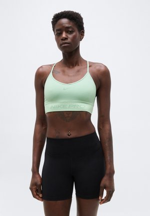 Atlétikus személy áll, világoszöld sportmelltartót és fekete testhezálló rövidnadrágot visel, egyszínű szürke háttér előtt.