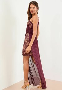 Frau in burgunderfarbigem, einseitigem Kleid mit Spitze und transparenter, langer Rock, trägt goldene Highheels und muschelförmige Ohrringe.