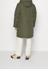 Olivgrön parkas med huva, åtdragen midja och långa ärmar. Bärs över vita byxor och ljusa skor med svarta och orange detaljer.