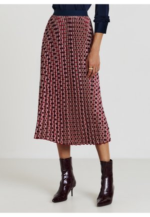 Femme portant une jupe midi plissée géométrique bordeaux et crème avec un chemisier bleu marine et des bottines bordeaux brillantes à talons.