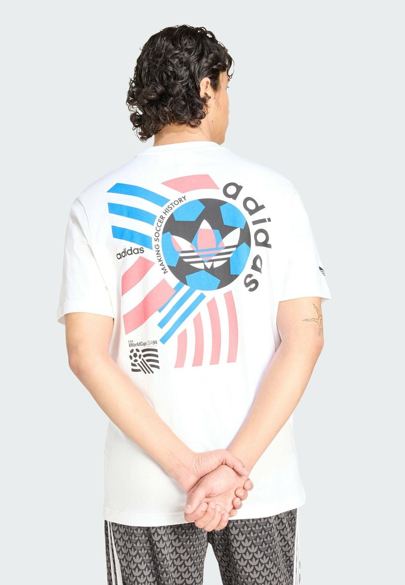 T-shirt en coton blanc avec imprimé graphique multicolore comprenant des éléments bleus, roses et noirs, dont un ballon de football et des rayures.