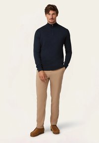 Marineblauer Reißverschlusspullover, beiger Slim-Fit-Hosen, braune Wildlederschuhe. Glatte Texturen, schlichtes Design, taillierte Form, minimalistische Details.