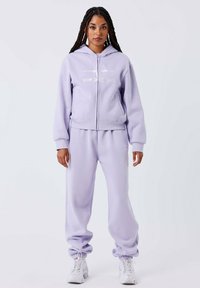 Lila zip-up hoodie en bijpassende joggingsbroek van zachte stof. De hoodie heeft een logo aan de voorkant, elastische manchetten en een jogger fit bij de enkels.