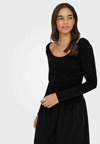 Sort langærmet top med bredt scoop-neck, lavet af et blødt stof, parret med en sort nederdel. Bemærkelsesværdige lagde sølv halskæder.
