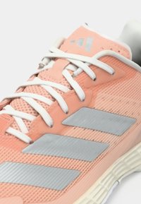 Brzoskwiniowy sportowy but z siateczki z białymi sznurowadłami i szarymi trzema paskami. Posiada teksturowane panele i amortyzowaną podeszwę.