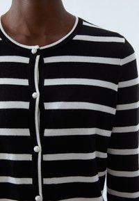 Černobílý pruhovaný kardigan s kulatým výstřihem, se zapínáním na perlové knoflíky a přiléhavým střihem. Měkká pletená textura.
