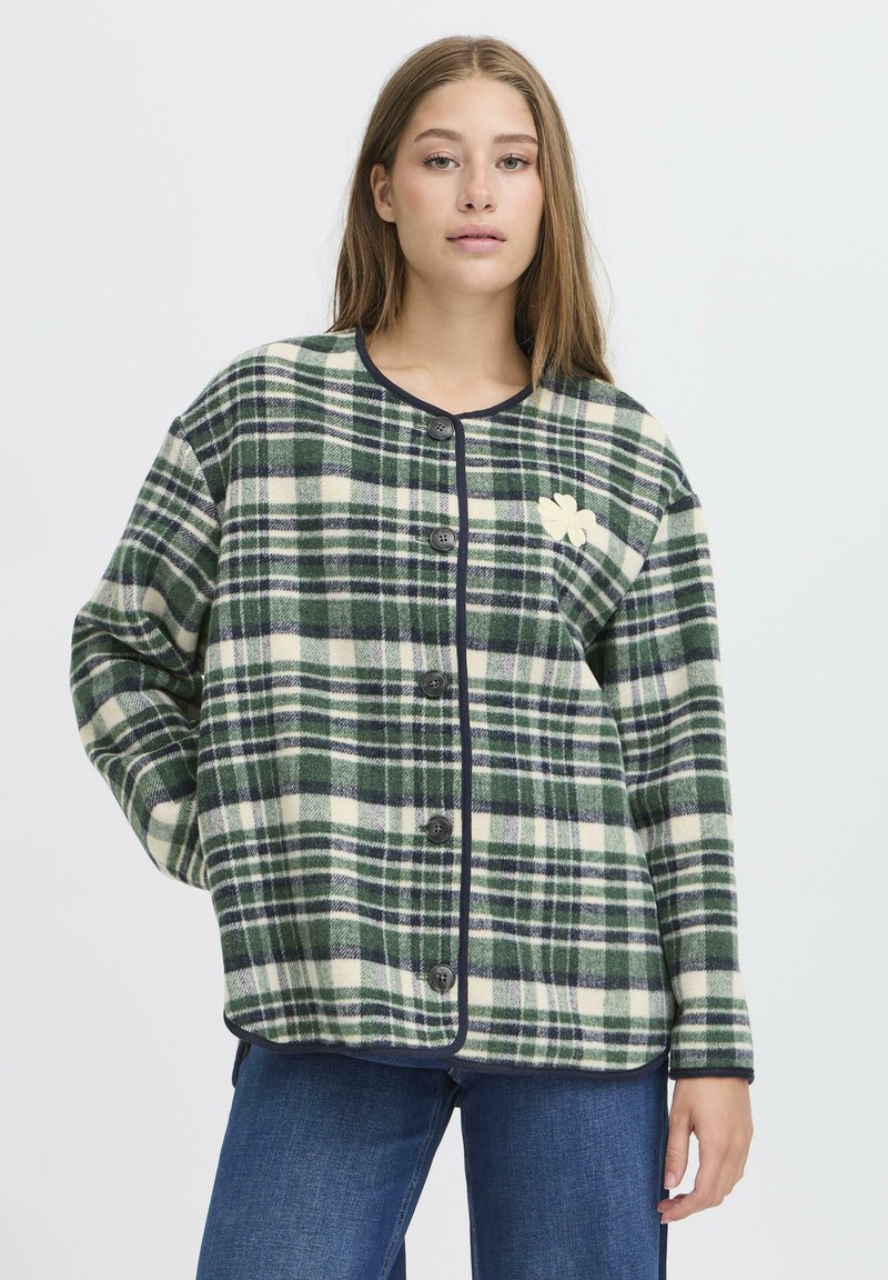 Veste à carreaux verte et crème avec des accents noirs, dotée d'un col arrondi, d'une fermeture à boutons et d'un petit motif brodé sur la poitrine.