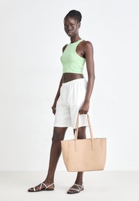 Sac fourre-tout beige avec deux longues anses, finition lisse, complété par un crop top vert clair et un short blanc à taille élastique. Sandales argentées.
