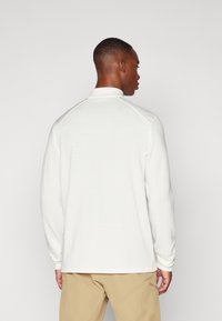 Chemise blanche à manches longues avec un design texturé en nervures et un col montant, portée avec un pantalon kaki, vue de dos.
