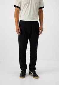 Mand med sorte bukser, sorte og hvide sneakers samt en hvid kortærmet skjorte med sorte ærmekanter, som står på et hvidt gulv.