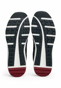 Suelas de zapatillas de goma negra con patrones texturizados; acento de media suela blanca. Detalle en el talón rojo visible, diseñado para tracción y comodidad.