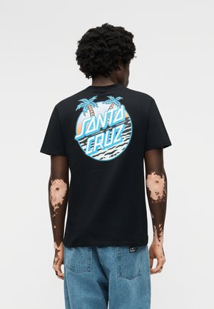 Άνδρας με λεύκη που φοράει μαύρο μπλουζάκι Santa Cruz με μπλε λογότυπο φοίνικα και κείμενο, συνδυασμένο με μπλε τζιν, γυρισμένος προς τα πίσω.