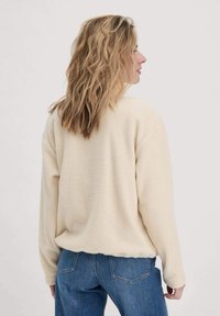 Frau mit welligem blonden Haar trägt eine cremefarbene Fleecejacke und blaue Jeans, steht seitlich mit größtenteils sichtbarem Rücken vor einem schlichten Hintergrund.