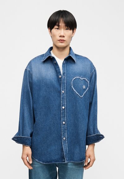 Jeune homme portant une chemise en denim ample aux manches retroussées, ornée d'un patch en forme de cœur effiloché sur la poitrine gauche.