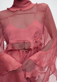 Διαφανής ροζ μπλούζα με βολάν στο ψηλό γιακά και μανίκια καμπάνα, φορεμένη πάνω από αντίστοιχη αμάνικη καμπίνα και φούστα.