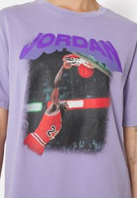 T-shirt en coton violet arborant un graphisme d'un joueur de basket dunkant, avec le texte "JORDAN" en violet foncé au-dessus de l'image.