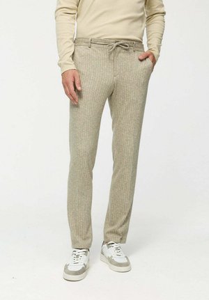 Beige texturierte Hose mit einem Kordelzugbund, die ein dezentes Muster aufweist. Kombiniert mit weißen und grauen Sneakers. Enganliegendes Design.