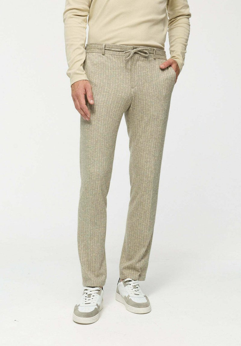 Beige textúrájú nadrág, állítható derékpánttal, diszkrét mintázattal. Fehér és szürke sportcipővel párosítva. Fitt szabású.