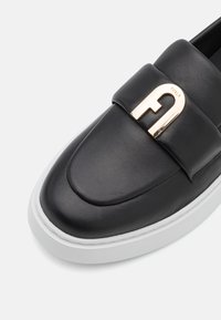 Zapato negro de cuero de tipo slip-on con parte superior acolchada, con un acento del logo de Furla en tono dorado y suela de goma blanca. Textura suave, forma redondeada.
