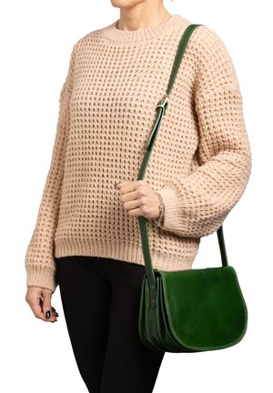 Persoon draagt een beige gebreide trui en zwarte broek, met een groene leren schoudertas met verstelbare band over de schouder.