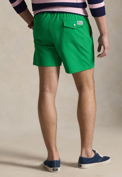 Πράσινα μαγιό βερμούδα με ελαστική μέση, μια πίσω τσέπη και ετικέτα. Συνδυασμένα με μπλε πάνινα παπούτσια. Ορατό δέρμα στα πόδια.