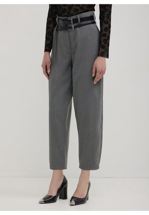 Personne portant un pantalon gris à fines rayures avec une double ceinture noire, un haut noir transparent à motifs floraux et des talons hauts noirs avec des bouts métalliques.
