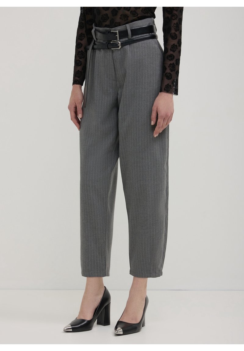 Personne portant un pantalon gris à fines rayures avec une double ceinture noire, un haut noir transparent à motifs floraux et des talons hauts noirs avec des bouts métalliques.