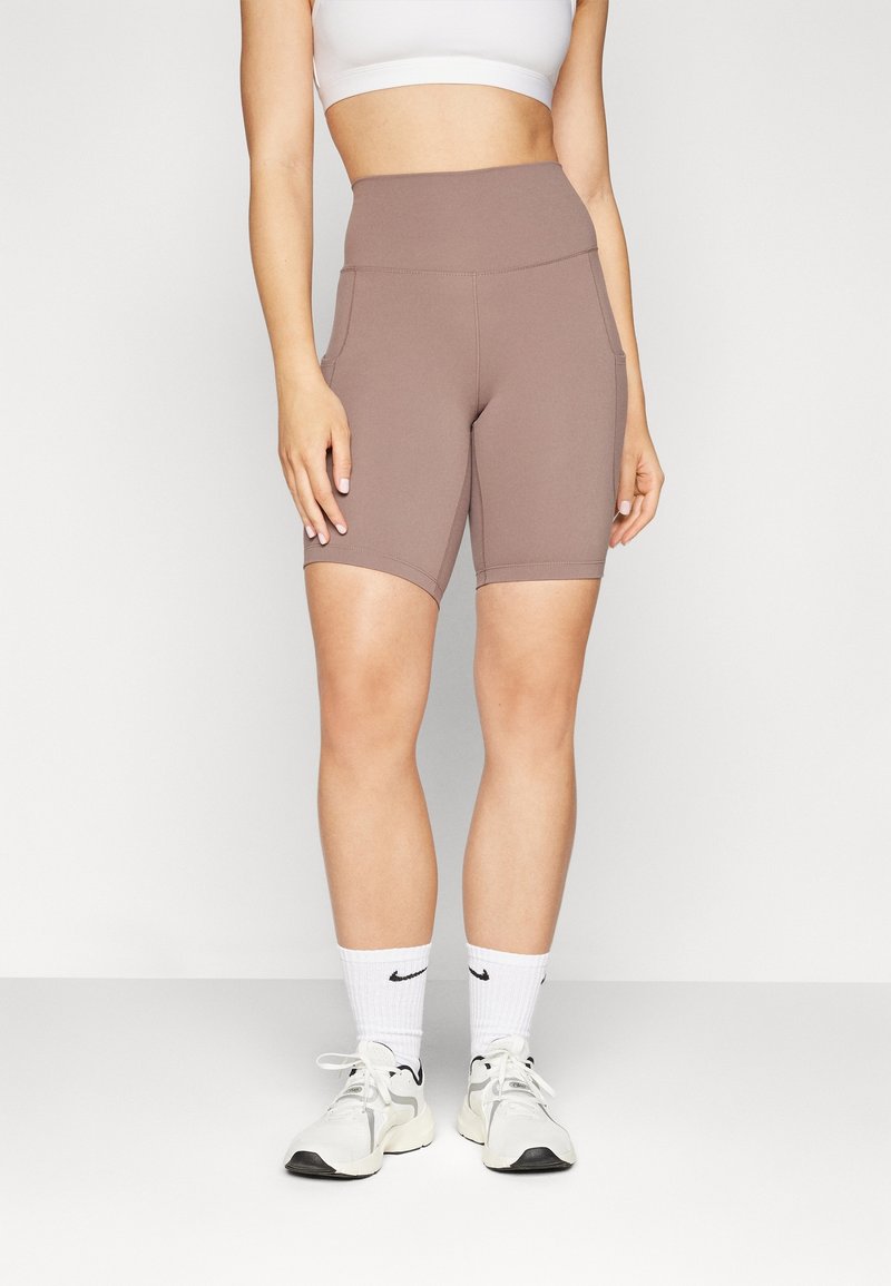 Femme portant un short de cycliste taille haute mauve, un soutien-gorge de sport blanc, des chaussettes Nike blanches et des chaussures de sport blanches, debout devant un fond uni.