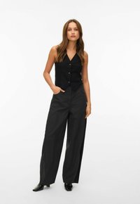 Žena stojí s jednou rukou v kapse, oblečená v černé bezrukávé vestičce na knoflíky, širokých černých kalhotách a černých botách na bílém pozadí.